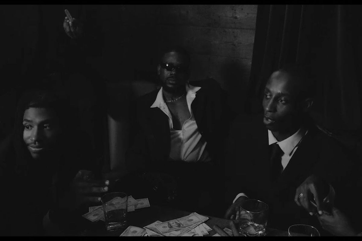 A black-and-white scene from Adekunle Gold's "Coco Money" music video, featuring men in suits seated around a table with cash, drinks, and cigars.