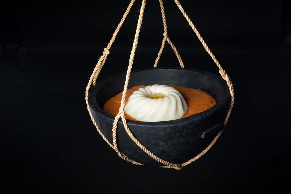 A black bowl is hanging in a net of fine ropes, filled with asida and an orange sauce.