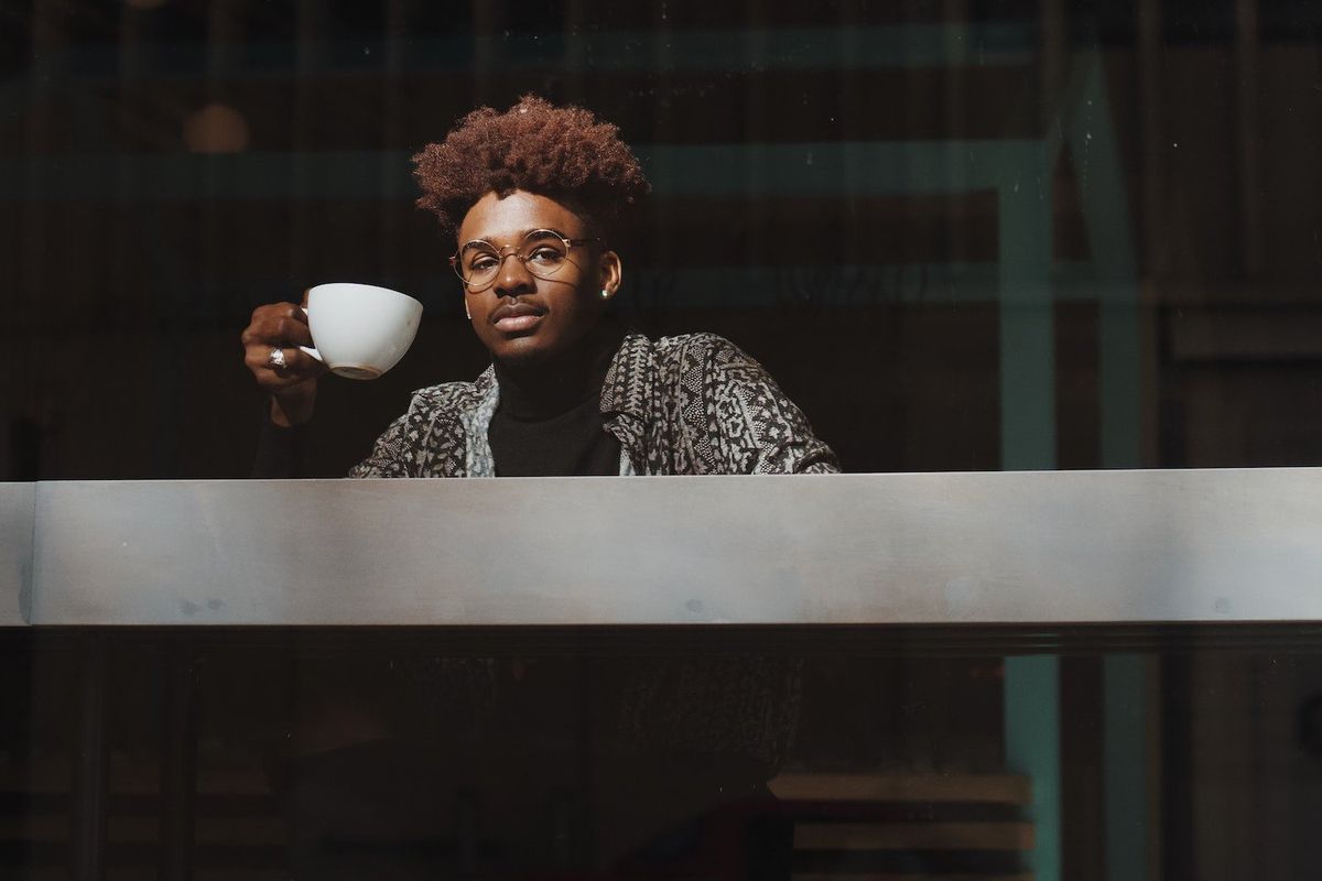 A Black man holds a mug and looks out the window.