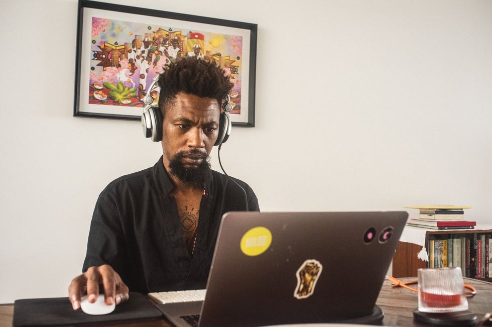A Black man with a short afro and goatee stits at his desk looking at an open laptop. 