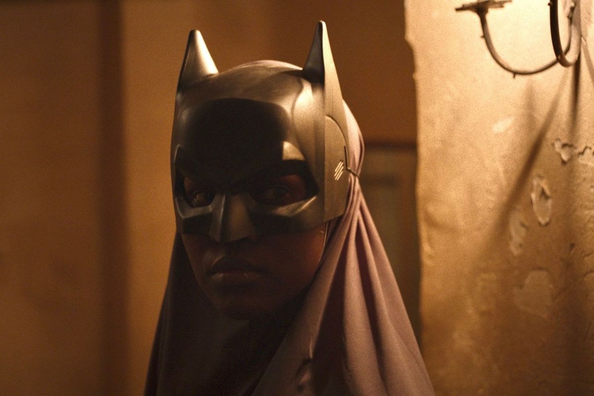 A Black woman wearing a grey veil and a superhero mask is standing in the dim light of a lamp.