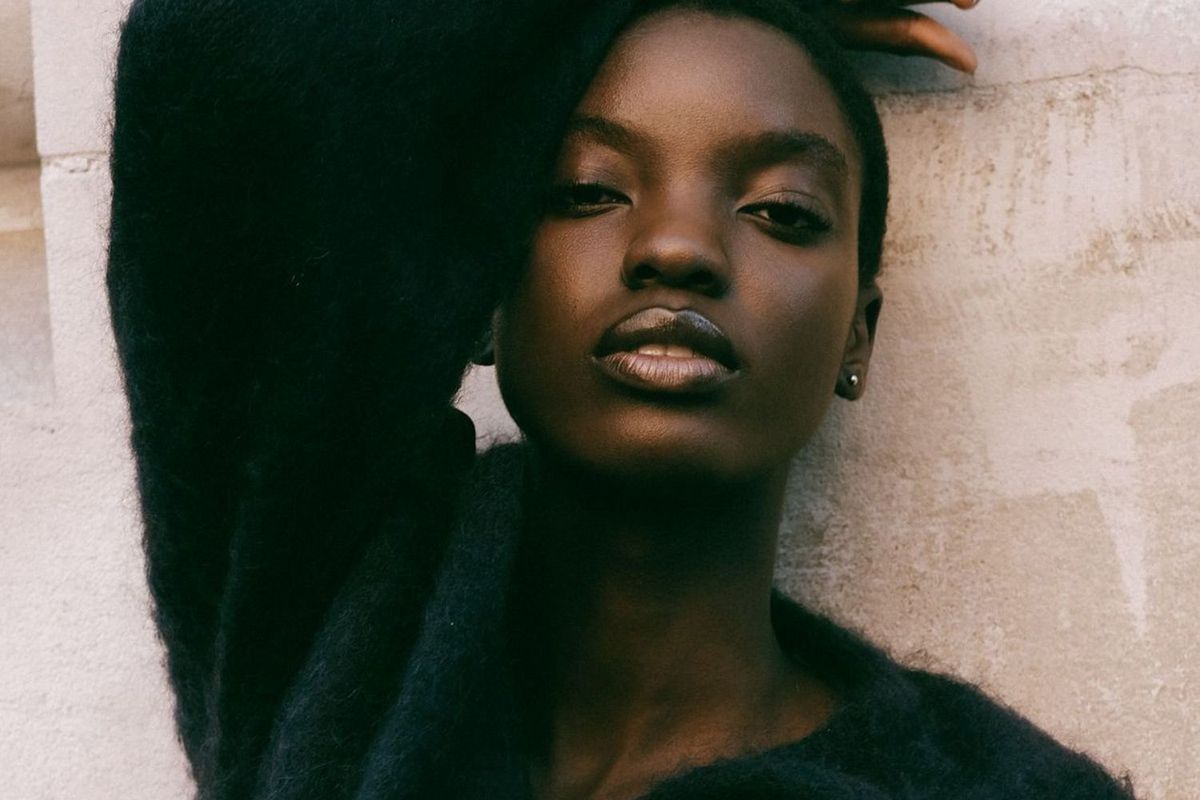 A close-up portrait of a woman wearing a dark textured top, leaning against a light wall with one arm raised over their head, gazing directly at the camera with a calm and confident expression.