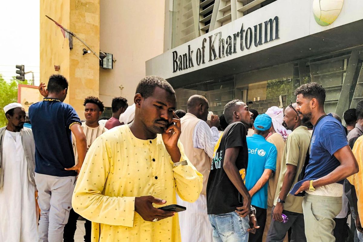 A concerned-looking man in a yellow jalabiya is holding his phone outside a Bank of Khartoum branch where several other men appear to be waiting with him.
