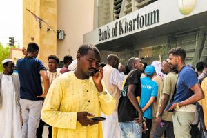 A concerned-looking man in a yellow jalabiya is holding his phone outside a Bank of Khartoum branch where several other men appear to be waiting with him. 