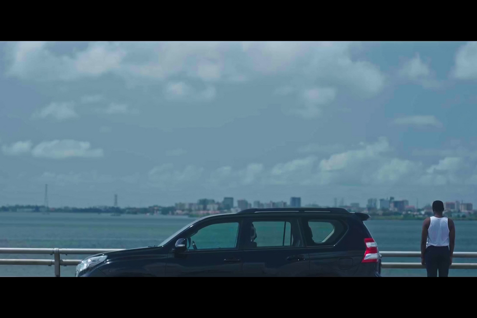 A film still from \u2018Over the Bridge\u2019 showing a man standing on a bridge and looking into the water, with an SUV beside him.
