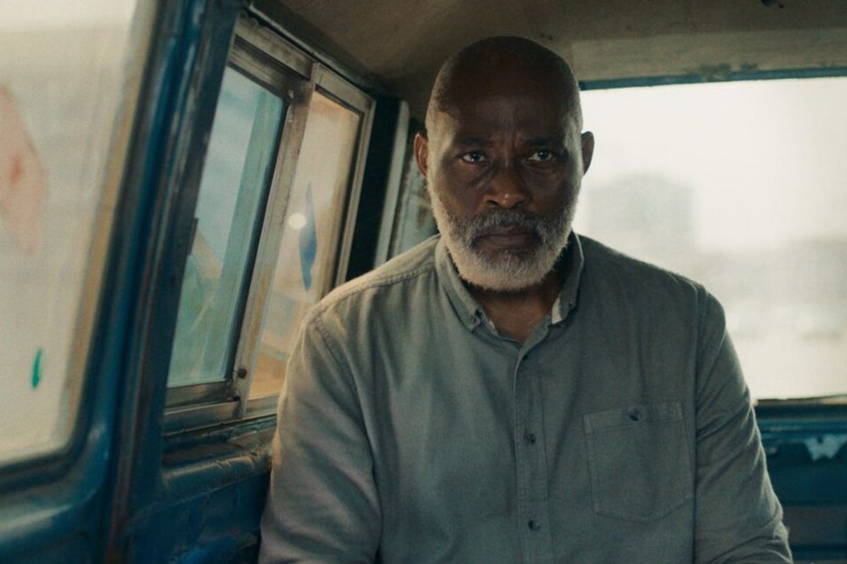 A film still of a man sitting in a van looking directly at the camera.