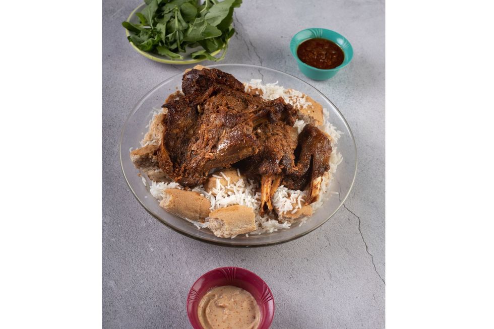 A glass plate filled with rice and soaked bread under slow-cooked lamb. Next to it on a grey table surface are three small bowls, two filled with dips and one with arugula.