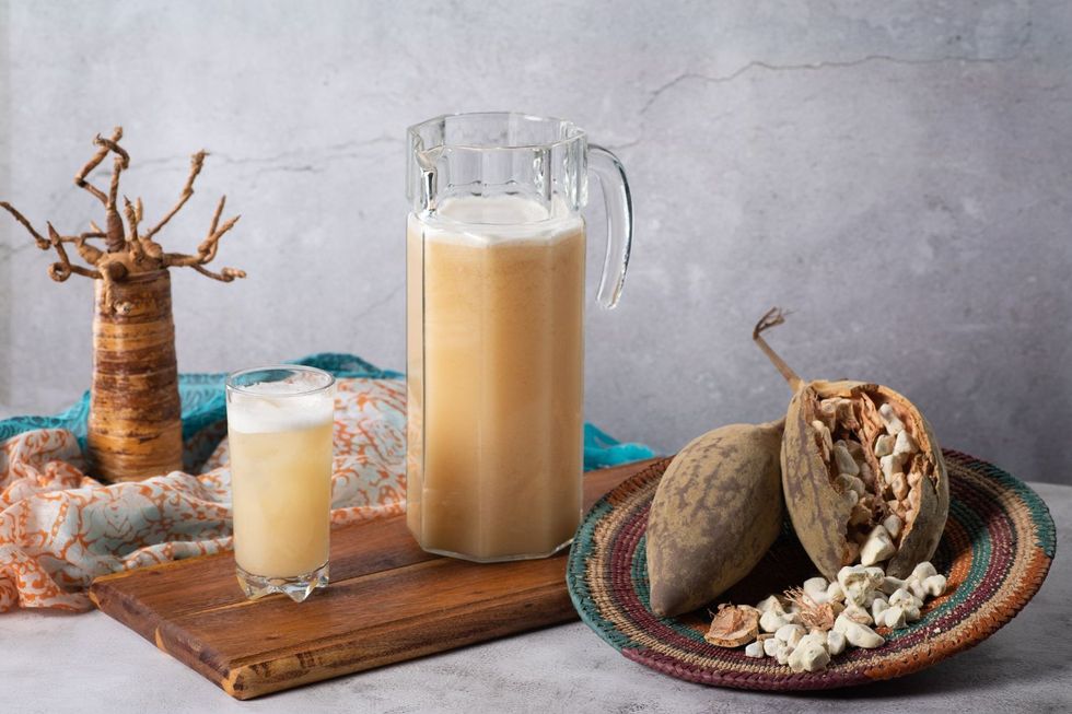 A jar and glass filled with yellow liquid are placed on a wooden board next to fruit arranged in a traditional Sudanese basket.
