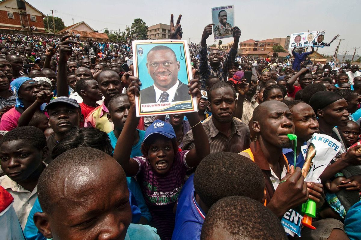 A large crowd of Ugandan supporters gather at a political rally, with several people holding up campaign posters of opposition leader Dr. Kizza Besigye. The mood is energetic, with people cheering, raising peace signs, and blowing whistles.