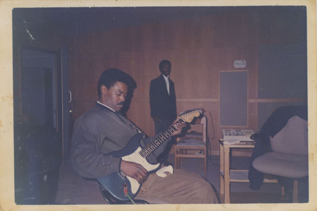 A man sitting in a wooden, dark studio playing a guitar. He is wearing beige trousers and a brown leather jacket. Behind him stands another man in a black suit, looking directly into the camera.
