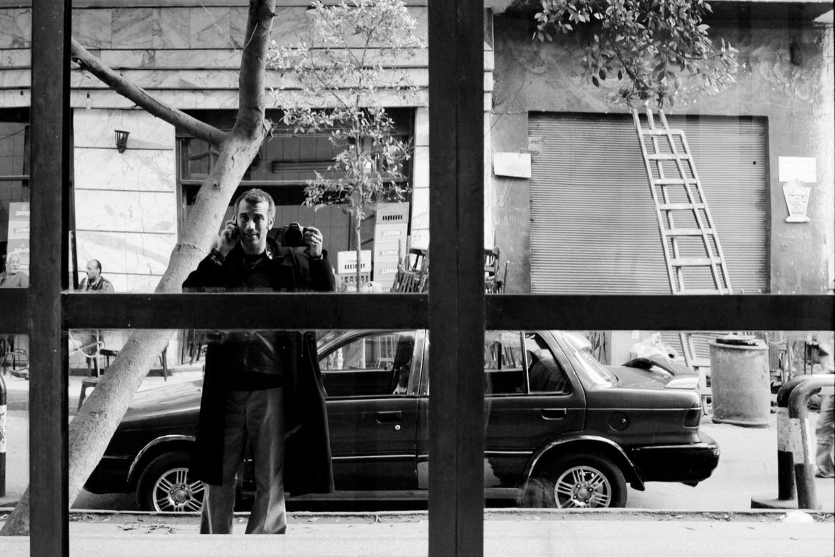 A man wearing a long dark coat is taking a picture of himself in the reflection of a door in the street.