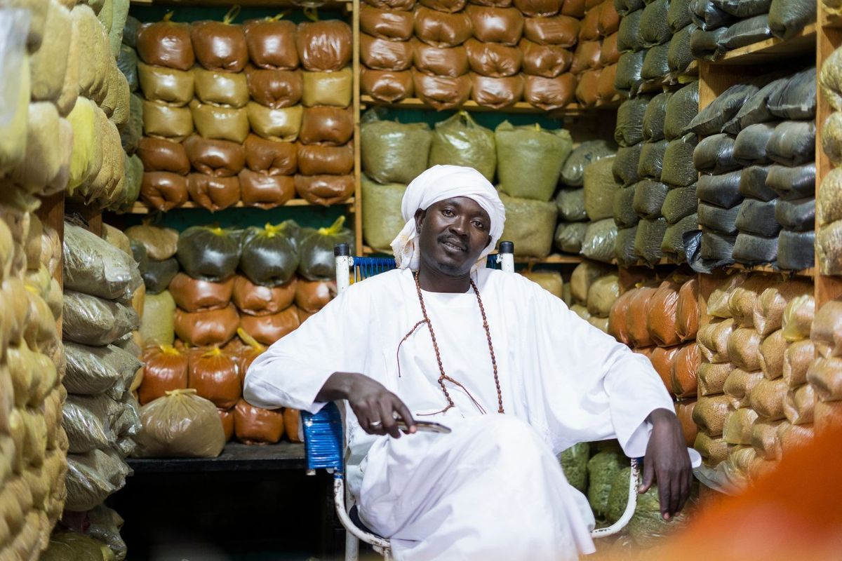 A man wearing a white jalabiya and turban is sitting in a small room. The walls are stacked with plastic bags that are filled with black, green, and orange colored grains.