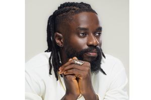 A man with a full beard and long locks styled in cornrows is pictured in a close-up portrait. He’s wearing a white shirt and a silver statement ring shaped like a panther. His hands are clasped together near his face, and he gazes thoughtfully into the distance. The background is a soft, neutral grey, keeping the focus on his expression.