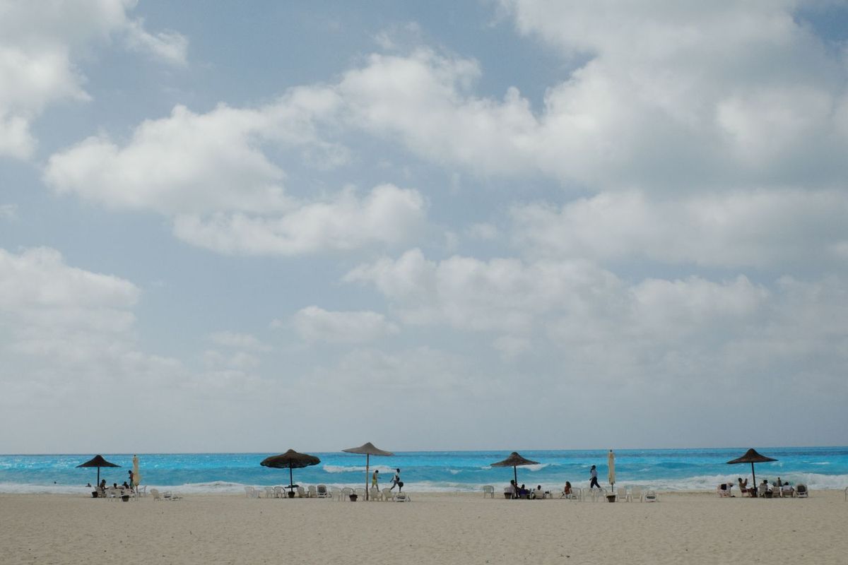 A Mediterranean shoreline with white umbrellas. Few people are walking on the beach.