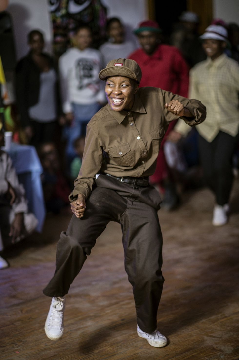 A pantsula dancer in action, wearing charcoal Dickies pants, an army green shirt and white All Stars. 