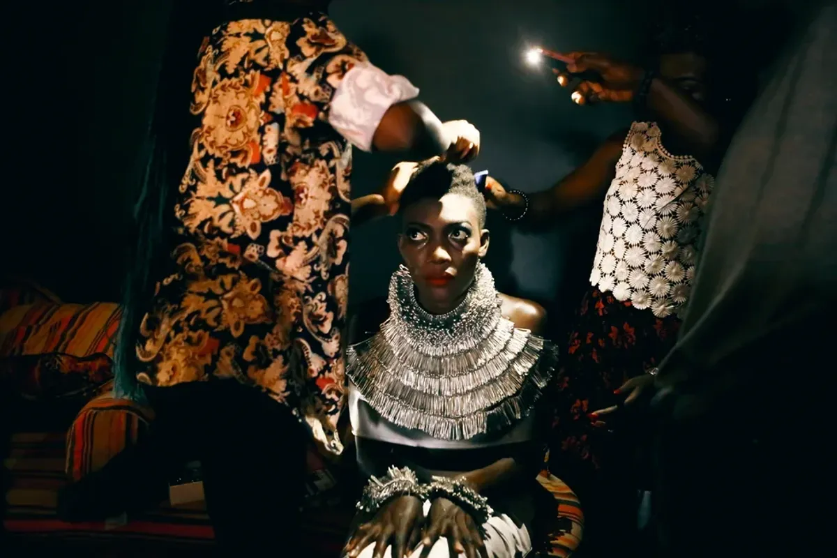 A photo of a woman sitting as two women make her hair in a room illuminated by a phone’s flashlight.
