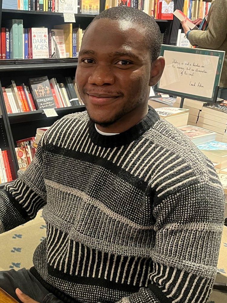 A photo of Chukwuebuka Ibeh sitting in a library with three copies of his book “Blessings” on the table.