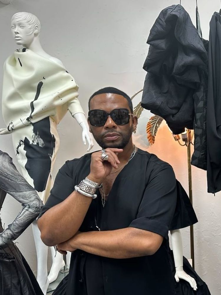 A photo of David Tlale in his New York showroom with some of his pieces on a hanger to his left.