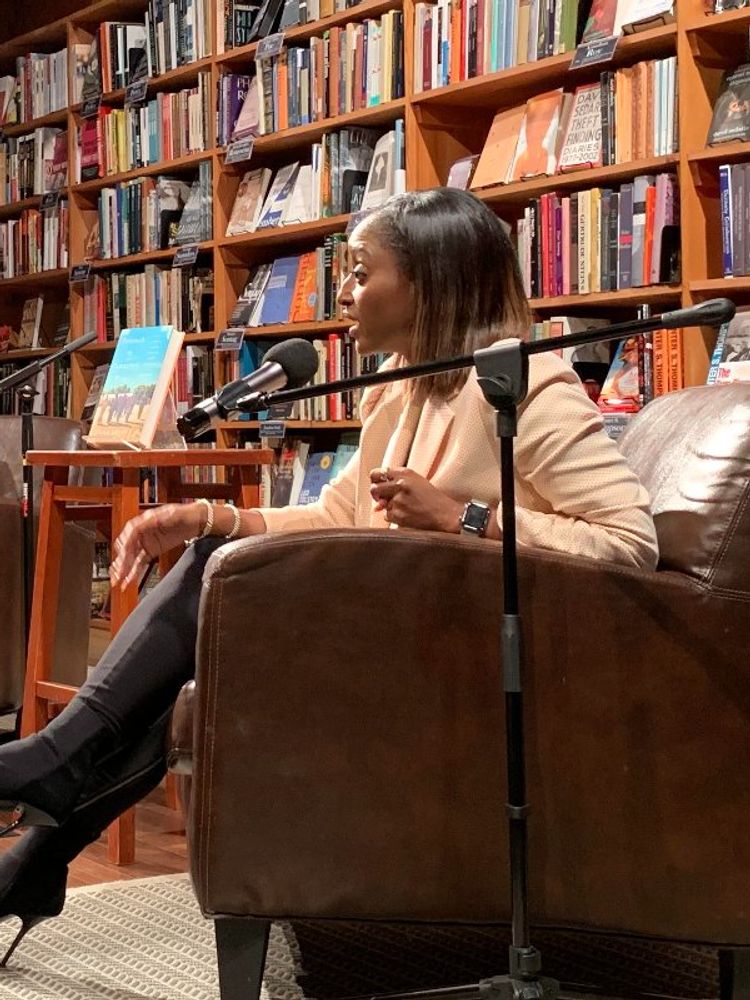 A photo of Patience Bulus (M) and OkayAfrica CEO, Isha Sesay (R) having a chat in a library, with Sesay’s book “Beneath the Tamarind Tree” on a stool in between them.