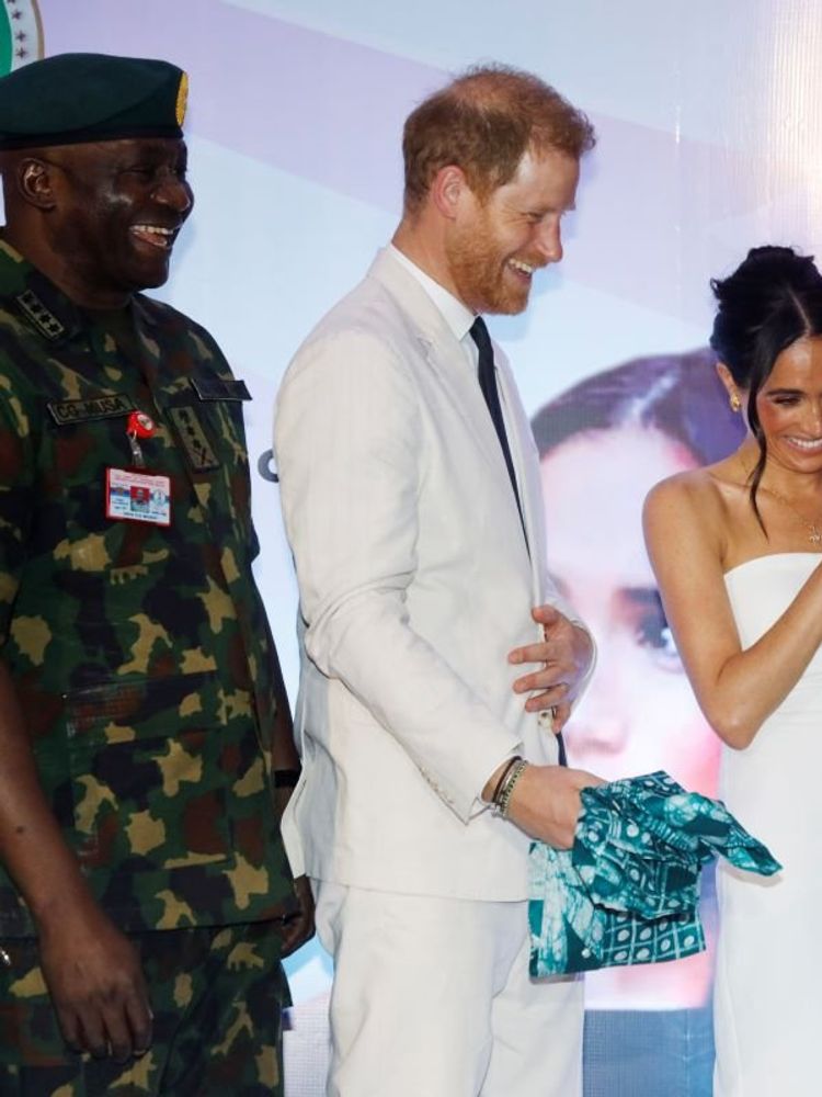 A photo of Prince Harry and Meghan Markle smiling as they receive outfits gifts from a woman.