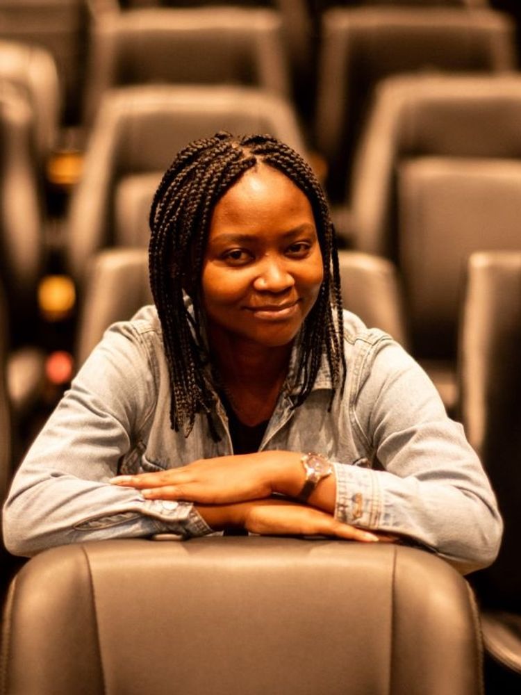 A photo of Sara Chitambo wearing a denim jacket and sitting, with her arms resting above each other on a chair in front of her.