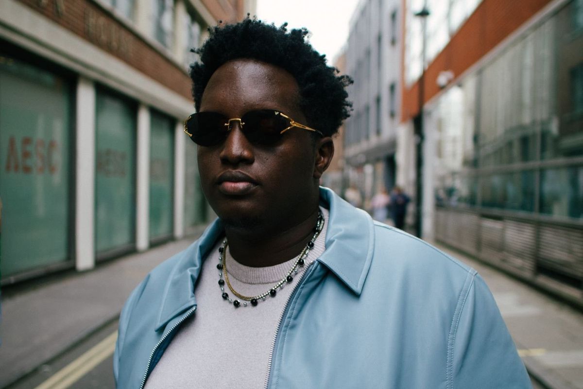 A portrait of Joshua Baraka wearing a light blue jacket and standing in a city street.