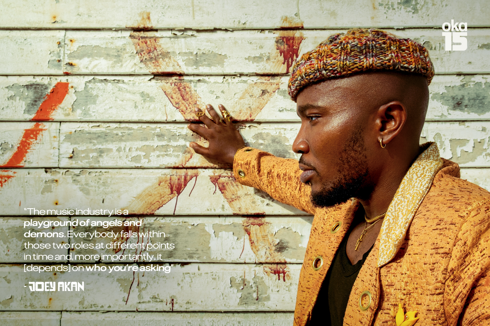 A profile shot of Nigerian music journalist Joey Akan wearing a yellow and gold suit and a beanie on his head, with his hand placed on a painted X on the wall.
