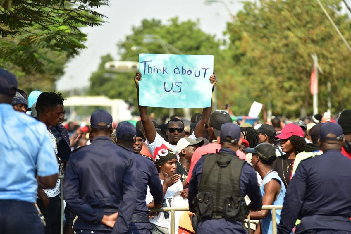 A protester holds a blue placard that reads, “think about us,” with other protesters around him and police officers in front of them.