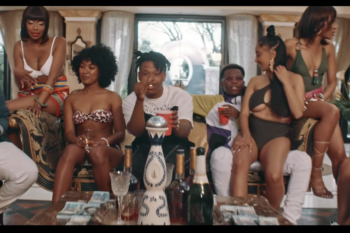 A screenshot from Nasty C's music video for 'Bookoo Bucks' showing him rapping surrounded by scantily dressed women and money. 