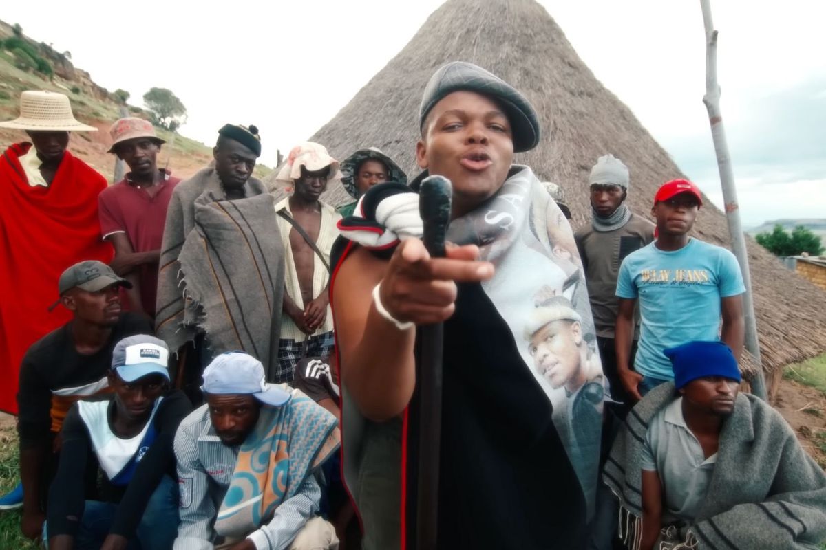 A screenshot of Sannere from the “Ke Holisitsoe Ka Malingoana” video. He stands in front of a hut, surrounded by people from the village, wearing a blanket he designed, and holding a fighting stick.