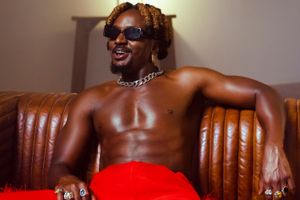 A shirtless Savara in red pants sit smiling on a leather couch