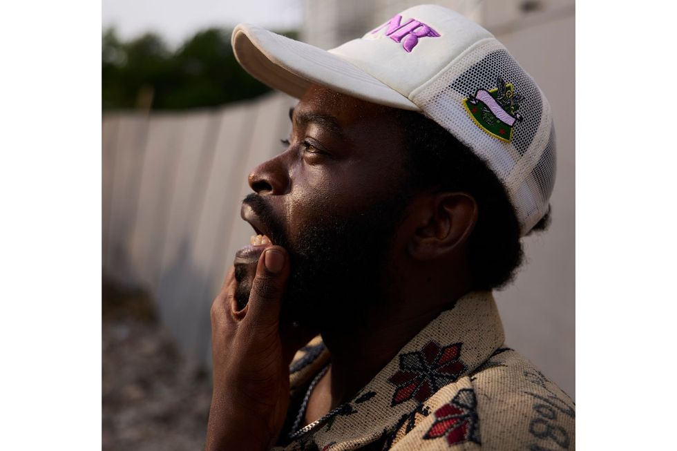 A side profile of Tim Lyre, who wears a white cap with pink writing, hand on chin, mouth open, and he looks off into the distance
