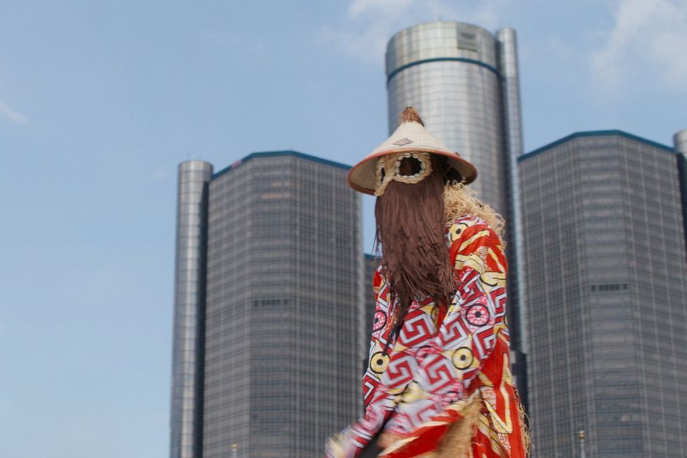 A still from the film of a man in a traditional Nigerian facemask standing in front of a building in Detroit.
