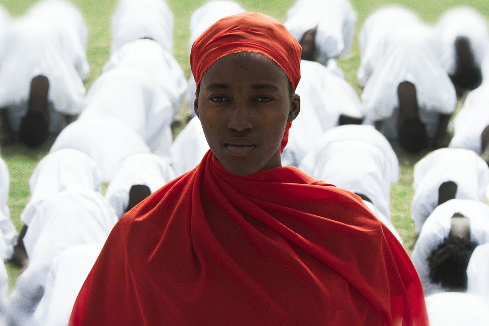 A still from the film of a woman in a headscarf looking at the camera, while people in white outfits bow in the background.