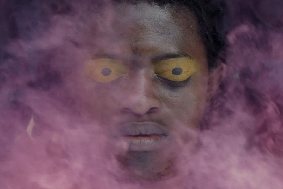 A still from the film, 'Omen,' of a pair of eyes with yellow eyeshadow on them covered by a cloud of pink smoke.