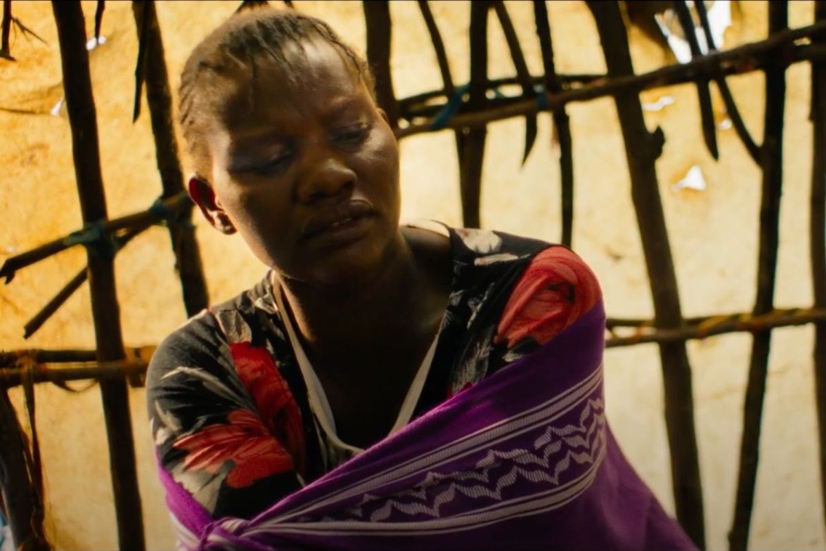 A still from the film 'One Woman One Bra,' showing a woman with braided hair, wearing a floral-patterned top and draped in a purple cloth, and sitting inside a wooden hut.
