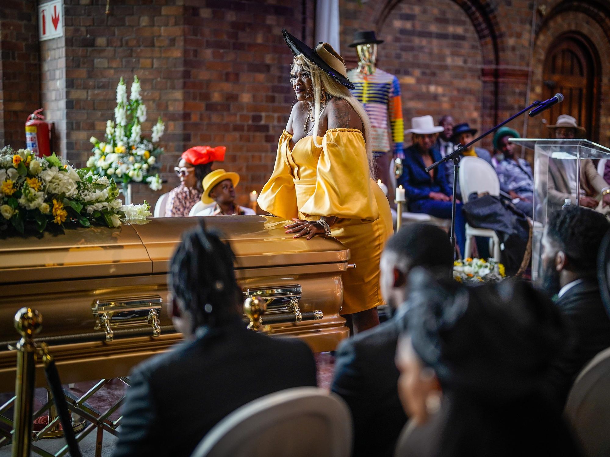 A still from the TV series, of a woman dressed in yellow standing at a coffin.