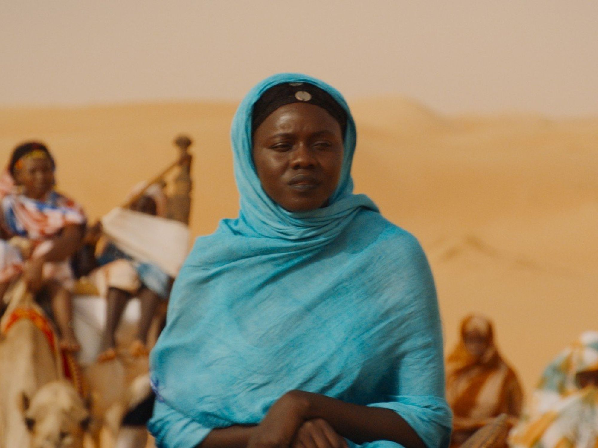 A still image from the film of a woman in the desert in a full headscarf looking pensive. 