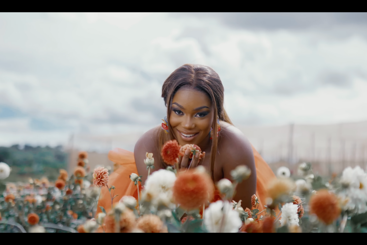 A still of Tamy Moyo in the visualizer for her latest single. She is in a garden of roses, gazing straight at the camera while wearing an orange dress.