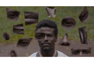 A Sudanese man wearing a white jalabiya and a beige tie is standing in a forest, looking directly at the camera. Parts of his face have been replicated and collaged all over the picture. 