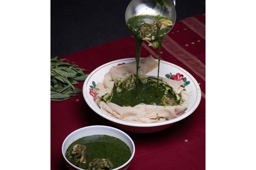 A white plate with kisra bread and molokhia with meat in an extra bowl. The molokhia is being poured onto the kisra.