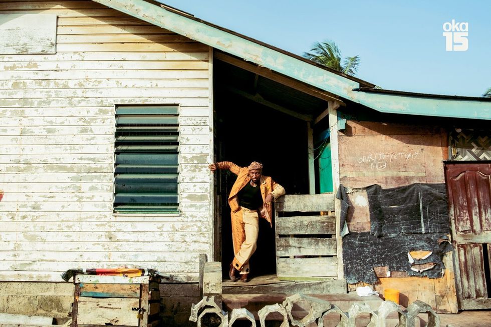 A wide shot of Afrobeats Intelligence Joey Akan standing in the entryway of a home. He is standing with his arms leaning on the walls on both sides, wearing a yellow and gold suit and a beanie on his head.