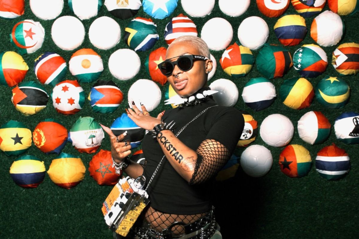 A woman poses in front of a vibrant backdrop made of balls decorated with African national flags. She's wearing a black top with black sunglasses and has a bold 'ROCKSTAR' tattoo on her arm.