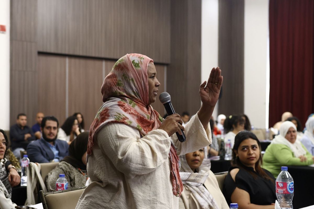 A woman wearing a beige and pink hijab is addressing people in a conference room, speaking into a microphone and gesticulating with a lifted arm.