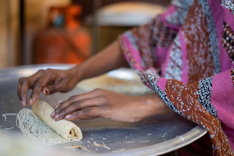 A woman wearing a purple toub is rolling kunafa malfufa on a silver tray.