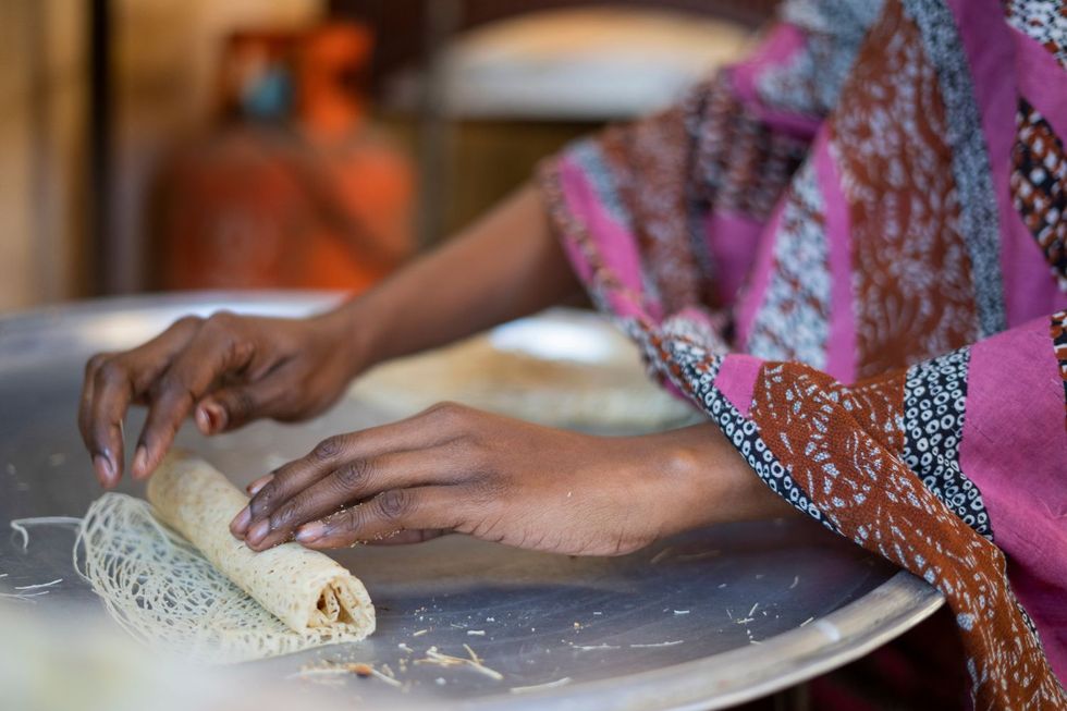 A woman wearing a purple toub is rolling kunafa malfufa on a silver tray.