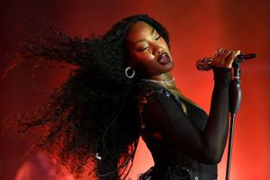 A woman with long curly hair performs passionately on stage under red lighting, holding a microphone and wearing hoop earrings and a sheer black outfit.