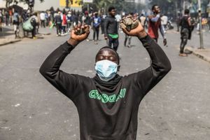 A young man with a mask partially covering his face kneels with his hands up, holding rocks, during protests against police brutality at the Central Business District in Nairobi, Kenya.