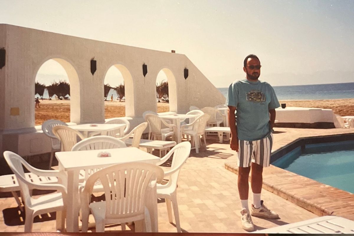 Ahmed Adel Wahby is standing by the pool in his Bawaki Beach Resort, wearing striped white and blue shorts and a blue t-shirt. Behind him is a white arc, around him white plastic tables with chairs, and in the distance, the Red Sea.