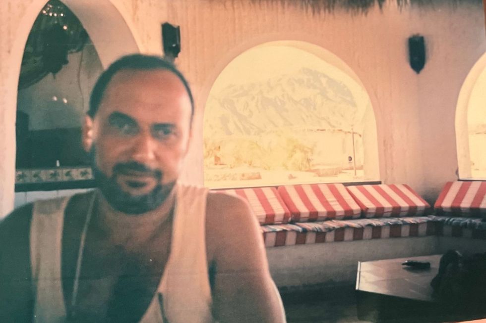 Ahmed Adil Wahby is sitting in the cafeteria, smiling into the camera and wearing a white tank top. Behind him is a large arched window through which one can see the Red Sea Mountains.